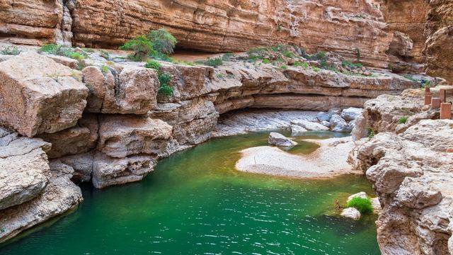 Smaragdgroene poel tussen gelaagde kalksteenwanden en rotsblokken in de kloof van Wadi Shab